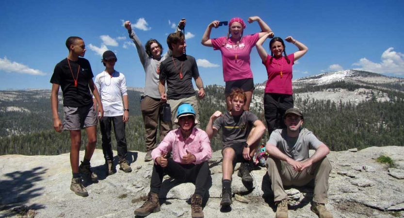 a group of people stand on a rocky overlook. Many of them are flexing their muscles. There are mountains in the background. 